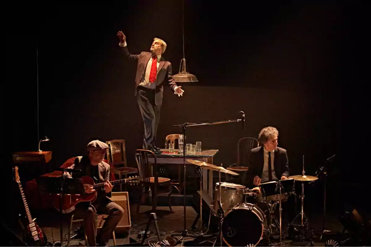 Tres músicos en un escenario oscuro, uno tocando la guitarra, otro la batería y un tercero de pie sobre una mesa con un micrófono.