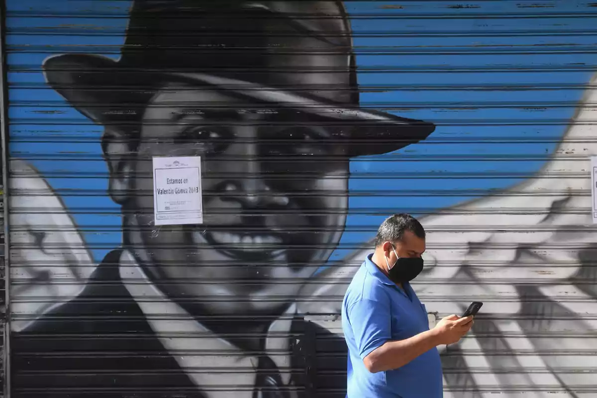 Un hombre con mascarilla y camisa azul revisa su teléfono frente a una puerta metálica con un mural de un hombre con sombrero y alas, y un cartel que dice "Estamos en Valentín Gómez 2643".