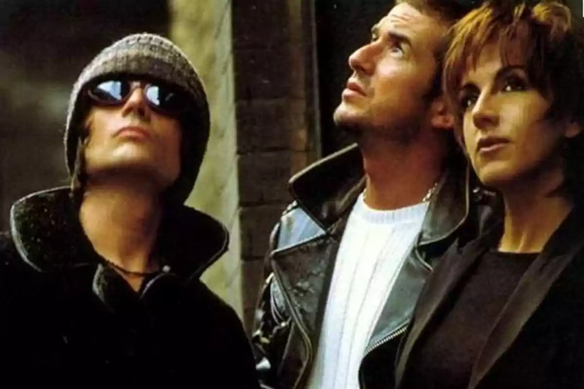 Tres personas mirando hacia arriba, una de ellas lleva gafas de sol y gorro, otra lleva una chaqueta de cuero y la tercera tiene el cabello corto.