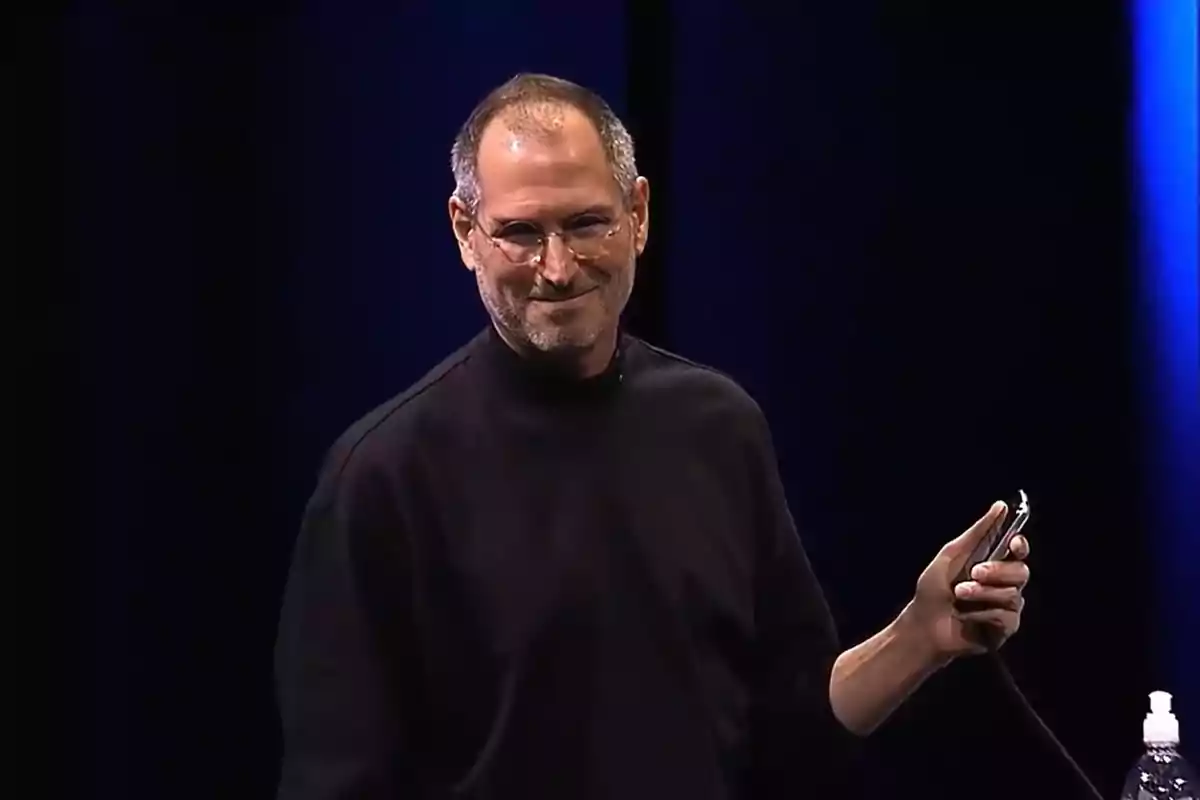 Persona con gafas y suéter negro sosteniendo un dispositivo en un escenario oscuro.