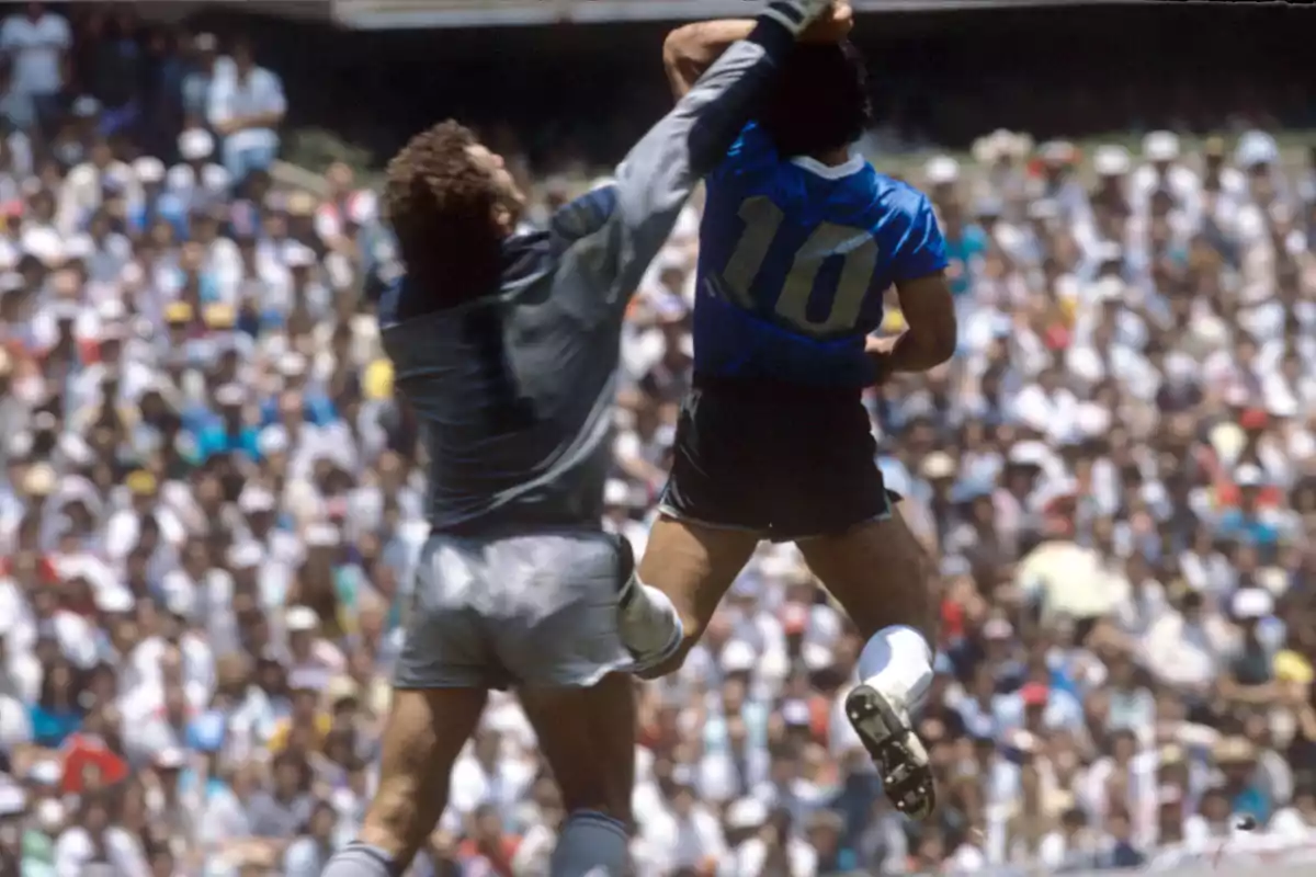 Un jugador de fútbol con la camiseta número 10 salta y extiende su brazo mientras un portero intenta detenerlo en un estadio lleno de espectadores.