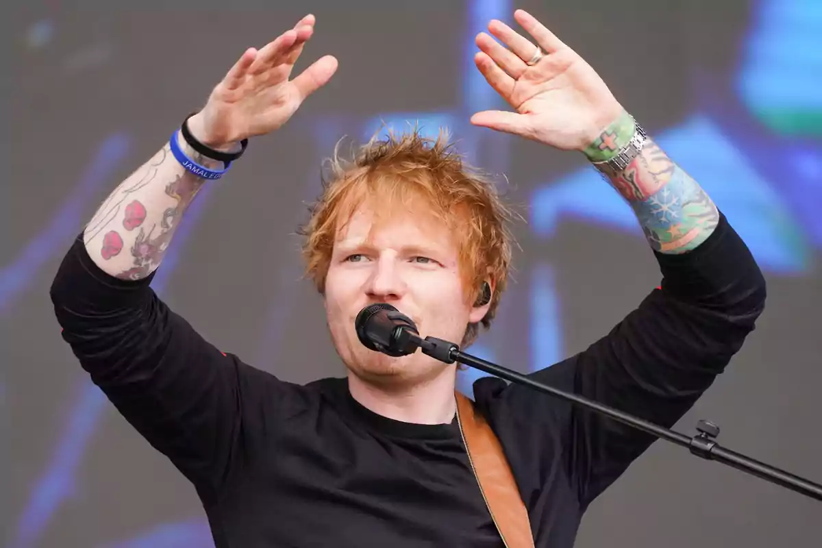 Persona con cabello pelirrojo y tatuajes en los brazos levantando las manos mientras canta en un micrófono.