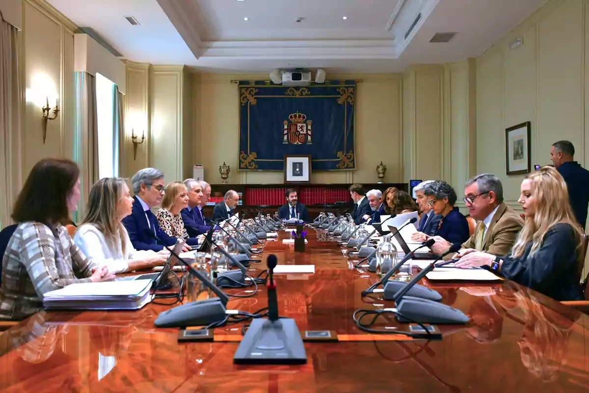 Personas sentadas alrededor de una mesa de conferencias en una sala de reuniones formal, con micrófonos y documentos frente a ellos, y un escudo de armas en la pared del fondo.