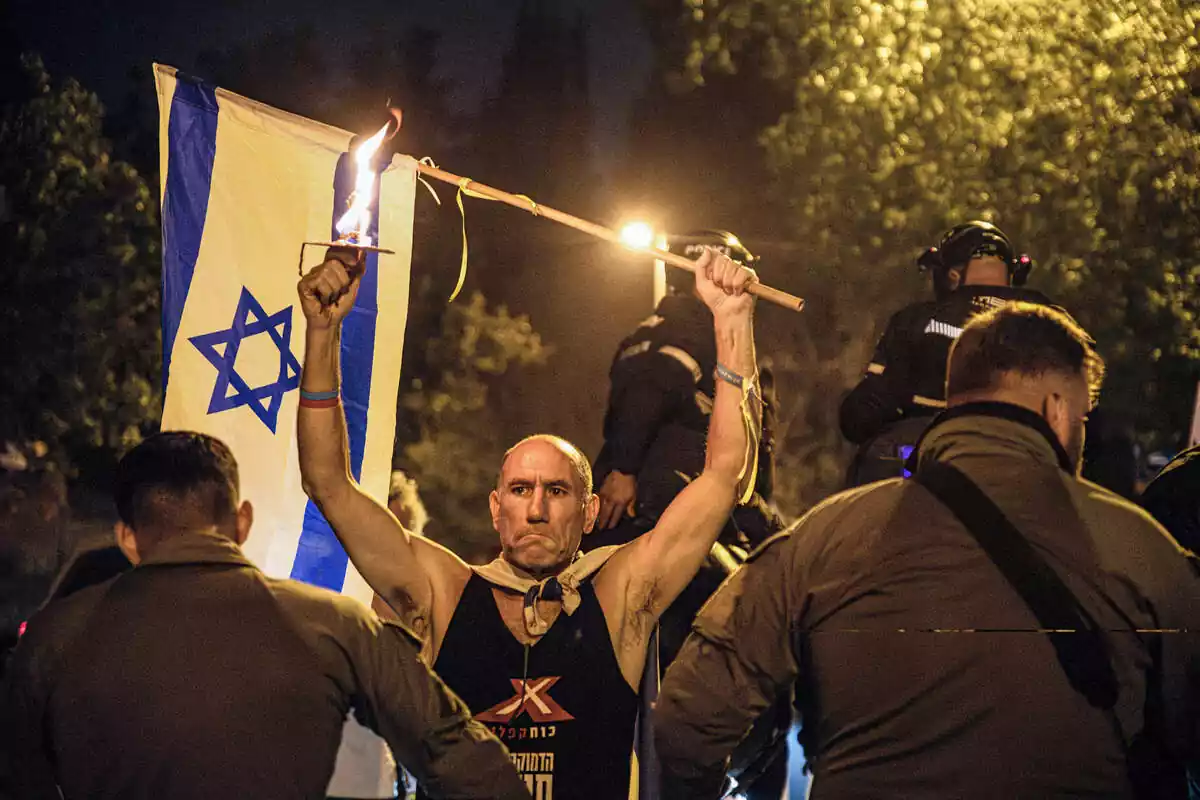 Un manifestante sostiene la bandera de Israel y una antorcha junto a una barrera policial alrededor de la residencia del primer ministro Netanyahu