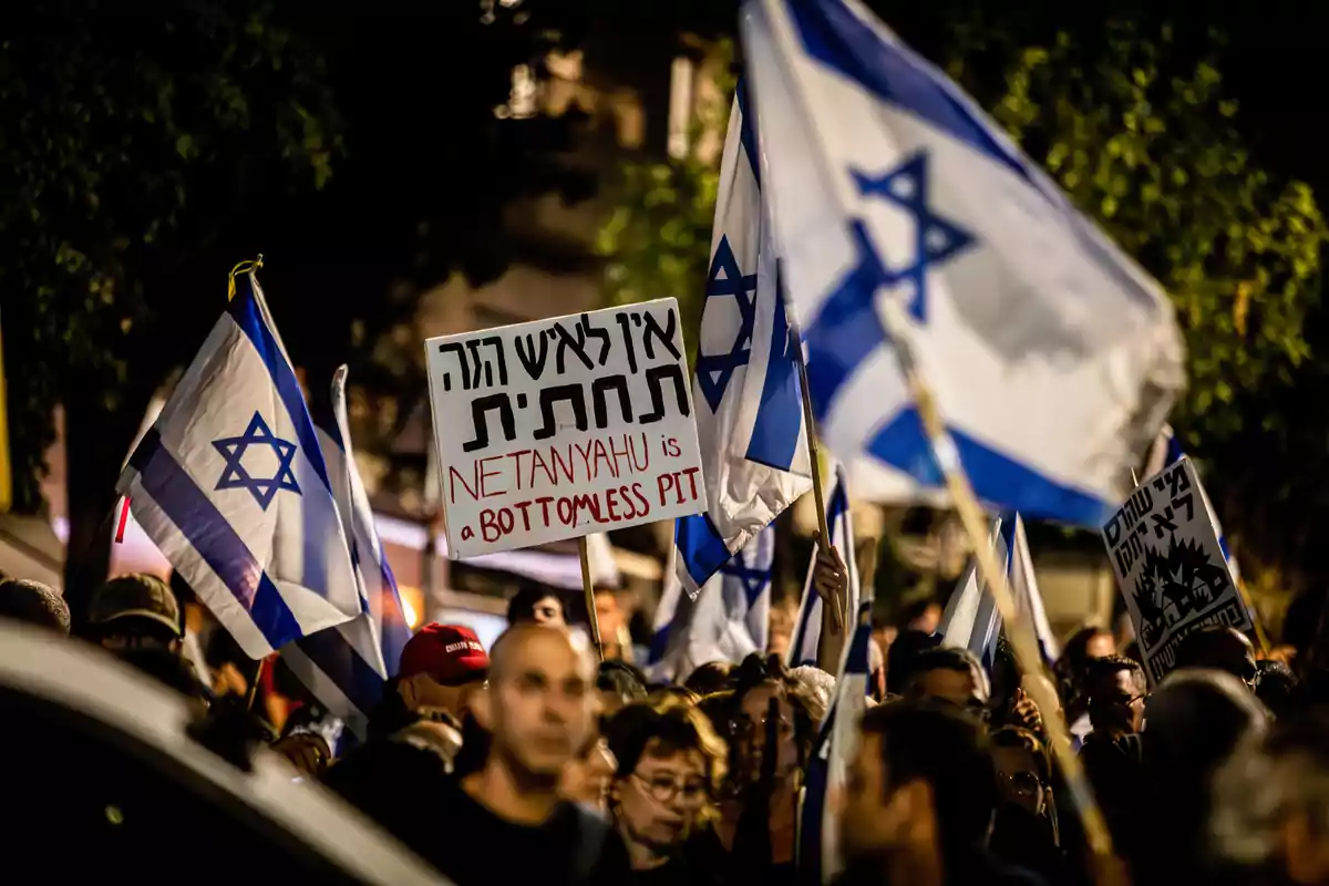 Una multitud de personas sostiene banderas de Israel y carteles de protesta, uno de los cuales dice "Netanyahu is a bottomless pit".