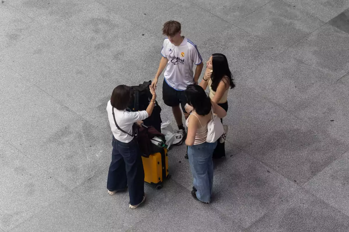 Un grupo de cuatro personas, tres mujeres y un hombre, están de pie en un espacio abierto de suelo gris, con una maleta amarilla y varias bolsas a su alrededor.