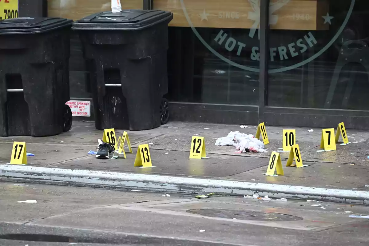Escena de un crimen con marcadores de evidencia numerados en la acera frente a un establecimiento, con dos contenedores de basura negros en el fondo.