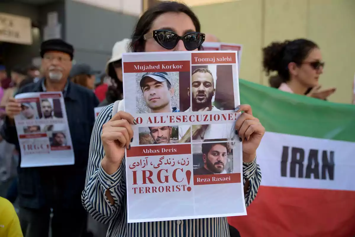 Una persona sostiene un cartel con fotos y nombres de varias personas, con un mensaje en italiano que dice "NO ALL'ESECUZIONE!" y otro mensaje en inglés que dice "IRGC TERRORIST!" mientras participa en una protesta; al fondo, otras personas también sostienen carteles y una bandera de Irán.
