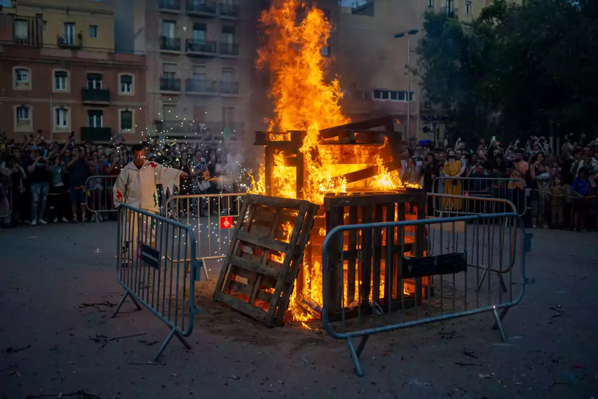 Una multitud observa mientras una gran hoguera de palets de madera arde intensamente en una plaza, con un hombre vestido de blanco avivando las llamas y chispas volando alrededor.