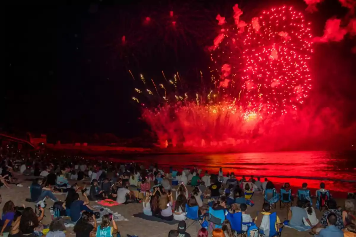 Una multitud de personas sentadas en la playa observando un espectáculo de fuegos artificiales rojos sobre el mar.