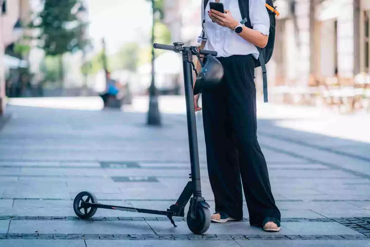 Persona de pie en la calle con un patinete eléctrico y usando un teléfono móvil.