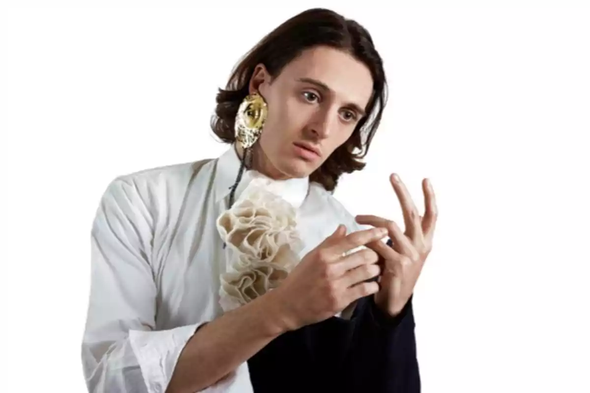 Un hombre con cabello largo y oscuro, vestido con una camisa blanca con un adorno de volantes en el pecho, lleva un pendiente grande y dorado en la oreja izquierda. Está mirando sus manos con una expresión pensativa. El fondo es blanco.